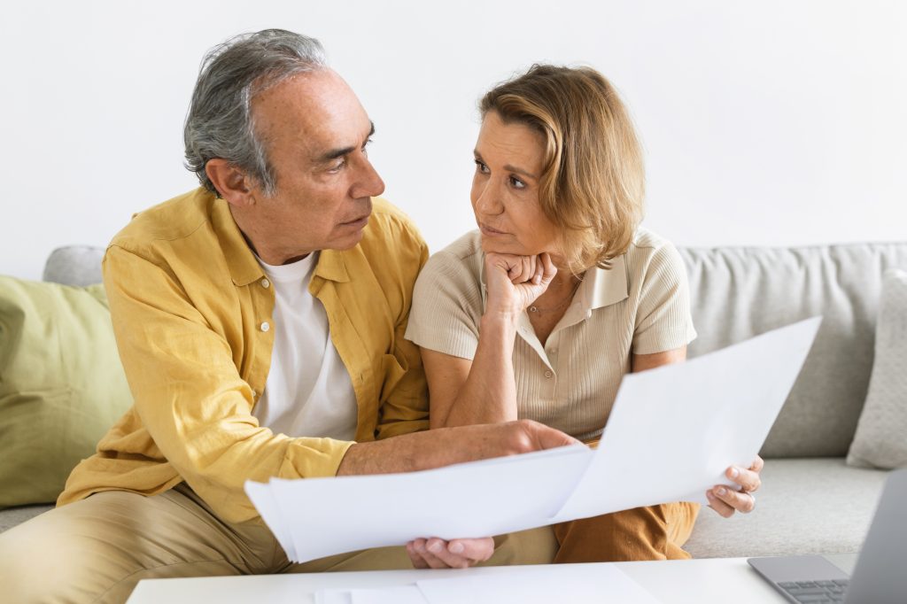 Concerned senior spouses sitting on sofa with papers, checking bills, making countings, working on